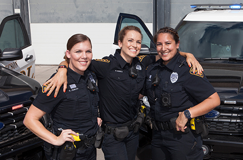 Three smiling female TPD patrol officers, in uniform, stand closely together, the middle one with arms around the shoulders of the other two. They are outside and their patrol vehicles, doors open, are behind them.